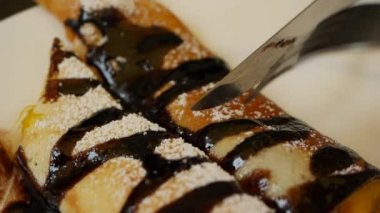 Çatal ve bıçak kullanan müşteri taze mango, çikolata ve pudra şekerli lezzetli krepler yiyor. Restoranda beyaz tabağa konmuş. Yakın plan.
