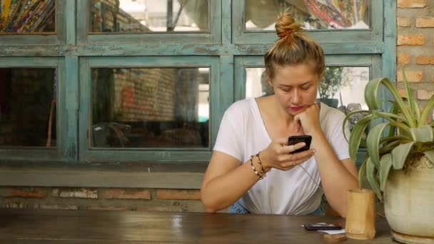 Menina pensativa usa um smartphone, escreve uma mensagem, conversando. Mulher à procura de informações no telefone e triste. Sentado no restaurante sozinho. Hora do pequeno almoço. Conceito de tecnologia e entretenimento — Vídeo de Stock