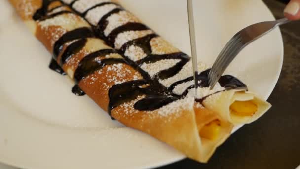 Le client qui utilise une fourchette et un couteau mange de délicieux rouleaux de crêpes avec de la mangue fraîche, du chocolat et du sucre en poudre placés sur une assiette blanche au restaurant. Gros plan — Video