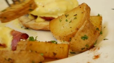 Benedict yumurtası yemek için çatal ve bıçak tutan kadın. Ekmek, domates ve tabakta kızarmış patates ile Benedict usulü yumurta. Brunch zamanı. Reklamcılık. Sağlıklı ketojenik yemek. Yakın plan.