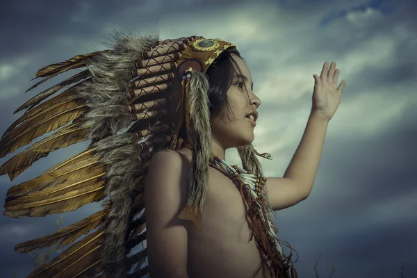 Child dressed as an American Indian at sunset, wears a plume of Indian feathers and breastplate of bones. he has raised his hand as a greeting to nature