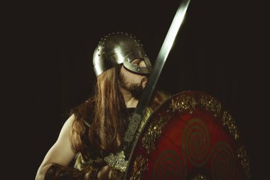 İskandinav, metalik miğferli ve hayvan derili Viking savaşçısı. deri eldivenler, altın süslemeleri ve çelik kılıç ile kırmızı ahşap kalkan