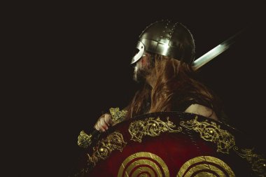 Metalik miğferli ve hayvan derili Viking savaşçısı. deri eldivenler, altın süslemeleri ve çelik kılıç ile kırmızı ahşap kalkan