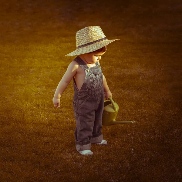 Autumn, little gardener in a garden of a house with a small green watering can watering the plants, wearing a cowboy breastplate and a straw hat