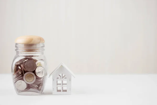 Money of savings coins in glass jar with model white house on wooden table. Property investment and house mortgage financial concept, Home protect, Insurance. With copy space for your text.