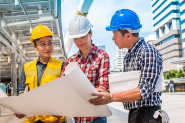 Üç endüstri mühendisi aşınma emniyet kask mühendislik çalışma ve çizimler muayene ile binanın önünde konuşurken. Mühendislik araçları ve inşaat kavramı.