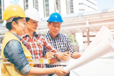 Üç endüstri mühendisi aşınma emniyet kask mühendislik çalışma ve çizimler muayene ile binanın önünde konuşurken. Mühendislik araçları ve inşaat kavramı.