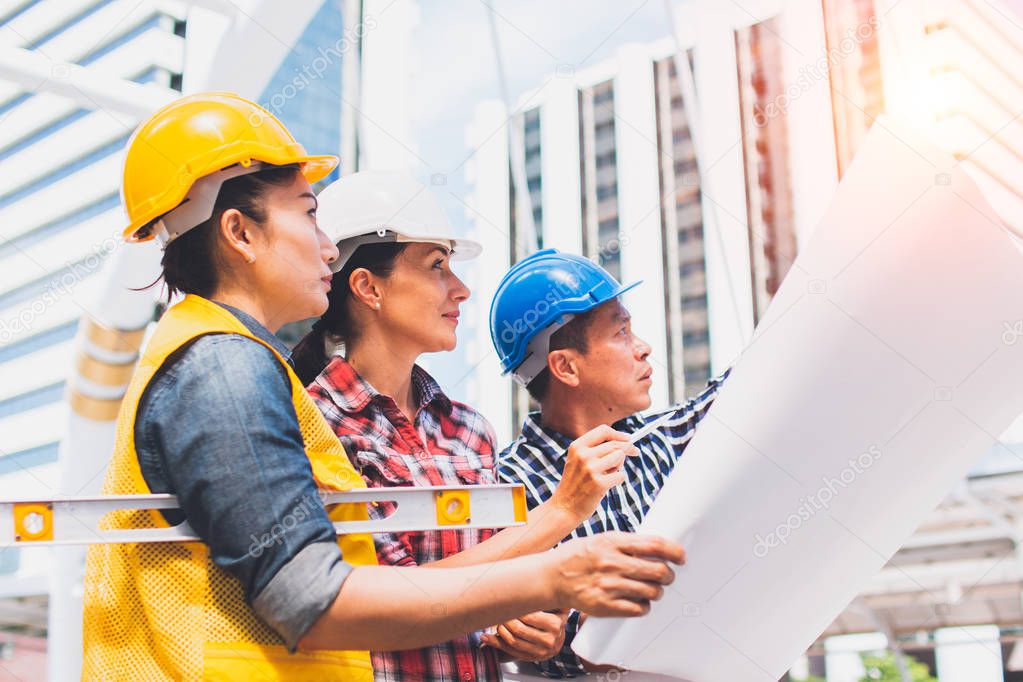 Three industrial engineer wear safety helmet engineering working and talking with drawings inspection on building outside. Engineering tools and construction concept.