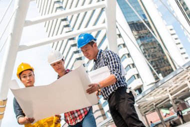 Üç endüstri mühendisi aşınma emniyet kask mühendislik çalışma ve çizimler muayene ile binanın önünde konuşurken. Mühendislik araçları ve inşaat kavramı.