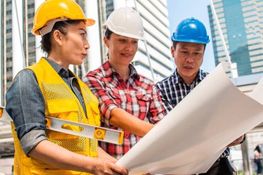 Üç endüstri mühendisi aşınma emniyet kask mühendislik çalışma ve çizimler muayene ile binanın önünde konuşurken. Mühendislik araçları ve inşaat kavramı.