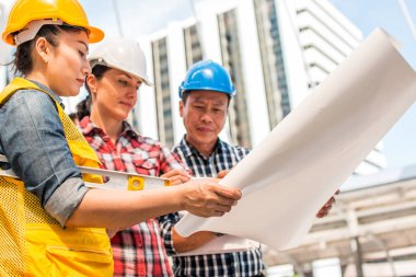 Üç endüstri mühendisi aşınma emniyet kask mühendislik çalışma ve çizimler muayene ile binanın önünde konuşurken. Mühendislik araçları ve inşaat kavramı.