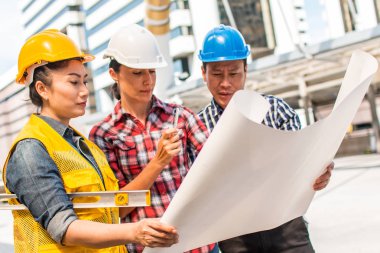 Üç endüstri mühendisi aşınma emniyet kask mühendislik çalışma ve çizimler muayene ile binanın önünde konuşurken. Mühendislik araçları ve inşaat kavramı.