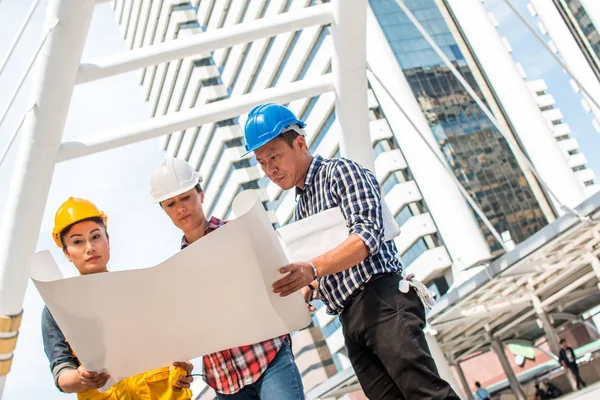 Üç endüstri mühendisi aşınma emniyet kask mühendislik çalışma ve çizimler muayene ile binanın önünde konuşurken. Mühendislik araçları ve inşaat kavramı.
