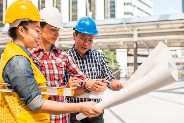 Üç endüstri mühendisi aşınma emniyet kask mühendislik çalışma ve çizimler muayene ile binanın önünde konuşurken. Mühendislik araçları ve inşaat kavramı.