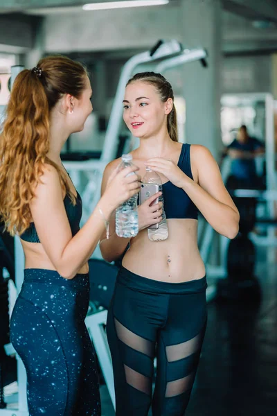 Fit beautiful young woman athletes in fitness clothes relaxing after workout in gym. Couple women standing holding water in bottle. Healthy, Sport, Lifestyle concept.