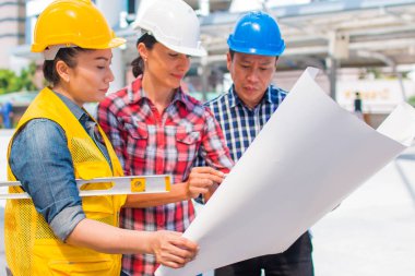 Üç endüstri mühendisi aşınma emniyet kask mühendislik çalışma ve çizimler muayene ile binanın önünde konuşurken. Mühendislik araçları ve inşaat kavramı.