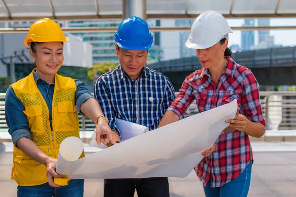 Üç endüstri mühendisi aşınma emniyet kask mühendislik çalışma ve çizimler muayene ile binanın önünde konuşurken. Mühendislik araçları ve inşaat kavramı.
