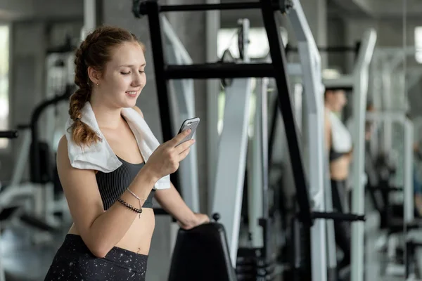 Happy Young attractive women fitness using smartphone resting in gym during morning exercise. Relaxation after hard workout in gym. Healthy sports lifestyle, Fitness concept. 스톡 이미지