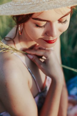 Hasır şapkalı bir kız bir çim üzerinde oturur, bir çiçek elleri, gözleri üzerinde ana odak tutar