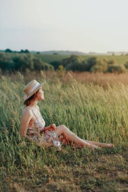 Hasır şapkalı bir genç kız çimenlerin üzerinde oturur, elinde bir sepet tarafında bir çiçek tutan