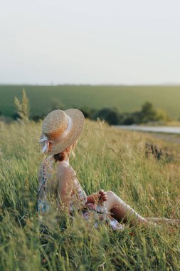 Hasır şapkalı bir genç kız çimenlerin üzerinde oturur, elinde bir sepet tarafında bir çiçek tutan
