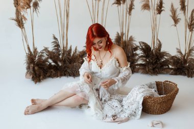 Düğün içeriği - stüdyoda beyaz bir arka plan üzerinde oturan ve süseşyaları ile elbisesini yapma genç bir güzel kızıl saçlı kız yakın çekim Portre. Kopyalama alanı