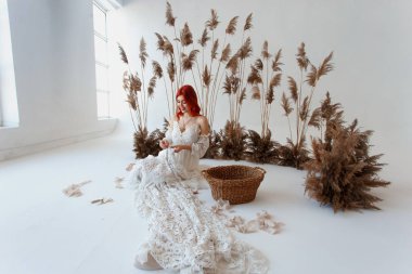 Düğün içeriği - stüdyoda beyaz bir arka plan üzerinde oturan ve süseşyaları ile elbisesini yapma genç bir güzel kızıl saçlı kız yakın çekim Portre. Kopyalama alanı