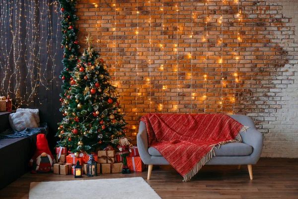 Kırmızı dekor topları ile Yeşil Noel ağacının Closeup, ve hediyeler kutuları, sarı çelenk ile bir tuğla duvara karşı, arka planda gri kanepe.