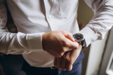 Evlenmeden önce bir erkeğin elini kelepçelemesine yakın çekim. Düğün detaycısı beyaz koluna mavi kol düğmesi takıyor. Saati ayarlayan beyaz gömlekli adam kol saatini ayarlıyor..