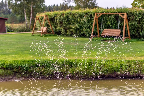 Two wooden swing benches in the garden with water drops in the foreground. Fountain water drops in the air with wooden garden swings in the background. Country summer view.