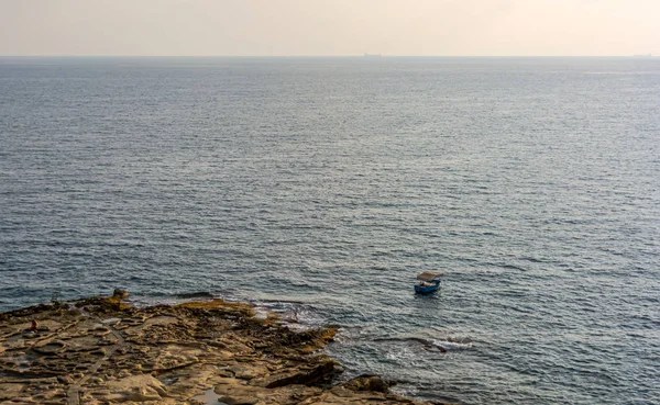 Lonely boat floating on the Mediterranean sea near Sliema coastline, Malta. Fishing boat is moored in the open sea near the Sliema shore and sandstone beach in the warm summer evening.