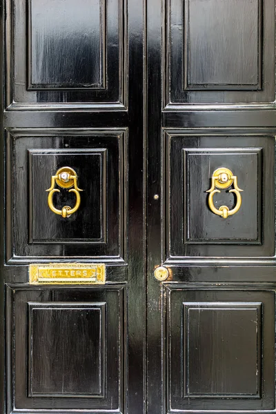 Black wooden door with golden vintage ornately shaped ring knockers and mail slot letterbox. Black door with golden hardware.