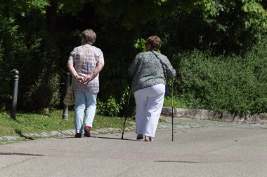 Haziran ayında Güney Almanya çok güneşli bir günde kıdem bayanlar ve baylar bir bankta oturan veya nordic walking sopalarla yürüyüş cloister Park görmek