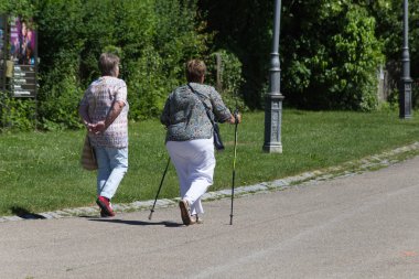 Haziran ayında Güney Almanya çok güneşli bir günde kıdem bayanlar ve baylar bir bankta oturan veya nordic walking sopalarla yürüyüş cloister Park görmek