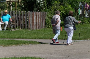Haziran ayında Güney Almanya çok güneşli bir günde kıdem bayanlar ve baylar bir bankta oturan veya nordic walking sopalarla yürüyüş cloister Park görmek