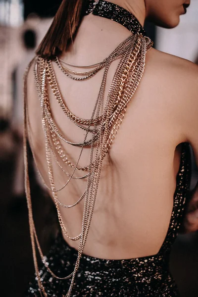 Gold and silver thin chains on a black dress on a young female model with burgundy lips standing back on backstage fashion week