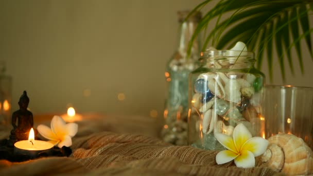 Glazen fles gevuld met schelpen, koralen, mariene onderdelen met kaarslicht, bloemen van de plumeria, Boeddha zittend voor decor — Stockvideo