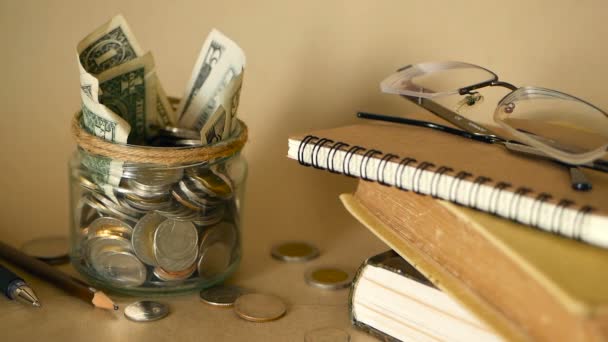 Boeken met glazen stuiver pot gevuld met munten en bankbiljetten. Collegegeld of onderwijs financiering concept. Beurs geld. — Stockvideo