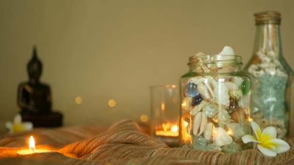 Стеклянная бутылка, наполненная ракушками, кораллами, морскими предметами со свечами, цветами плюмерии, сидящий Будда для декора — стоковое видео