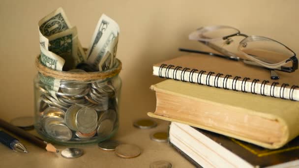 书里装满了硬币和钞票的玻璃便士罐子。学费或教育经费的概念。奖学金资金. — 图库视频影像