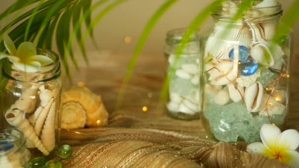 Vaso di vetro pieno di conchiglie, coralli, oggetti marini con luci bokeh, fiori di plumeria frangipani per arredamento — Video Stock