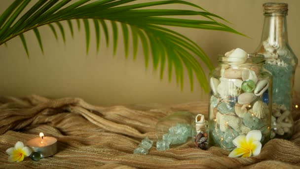 Glass bottle filled with seashells, corals, marine items with candle lights, plumeria frangipani flowers for decor — Stock Video