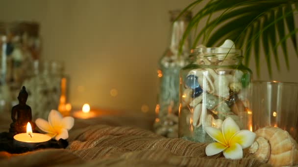 Glazen fles gevuld met schelpen, koralen, mariene onderdelen met kaarslicht, bloemen van de plumeria, Boeddha zittend voor decor — Stockvideo