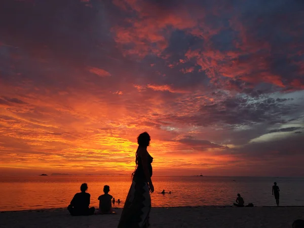 Silhouette of people on sunset beach. Side view of female silhouette on tropical coastline against multicolored tropical sunset sky with people around, Thailand
