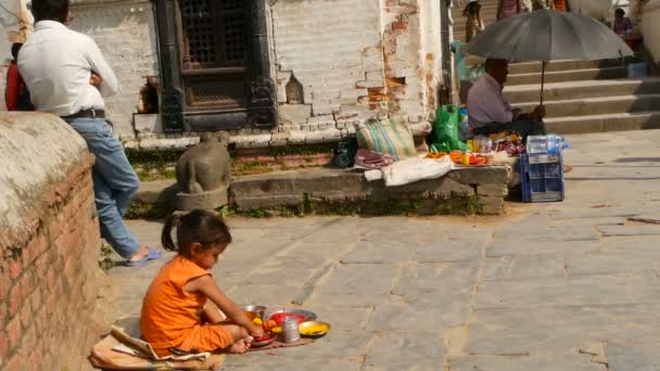 KATHMANDU, NEPAL - 8 EKİM 2018 Küçük etnik kız çeşitli dini özellikler satıyor, tapınağın taş kaldırımında oturuyor. Pashupatinath 'daki ghatlar. Geleneksel Hint geleneği. — Stok video