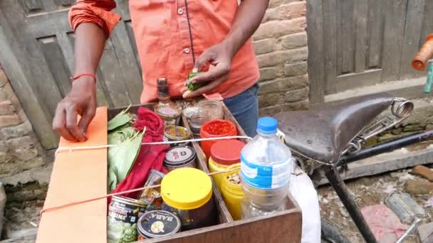 BHAKTAPUR, KATHMANDU, NEPAL - 18 de octubre de 2018 El hombre de las cosechas prepara hojas de betel en la calle. Hombre étnico sin rostro con caja de ingredientes en bicicleta preparando producto energizante con hojas verdes. — Vídeos de Stock