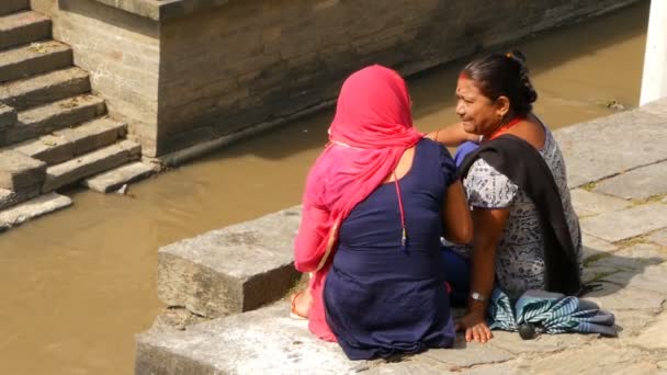 KATHMANDU,NEPAL - 2018年10月12日.石の街の堤防の女性。少数民族の女性が座って、太陽の下で汚れた浅い都市川の石の端に話しています。パシュパティナート・ヒンドゥー教寺院付近の火葬. — ストック動画