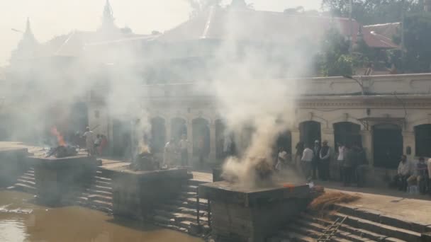 KATHMANDU, NEPAL 12 OCTOMBRIE 2018. Oamenii stând pe ghats arzând. Vedere a ghats de piatră cu paie arzătoare și lemn de foc în ritual de incinerare lângă Templul Hindus Pashupatinath în lumina soarelui — Videoclip de stoc