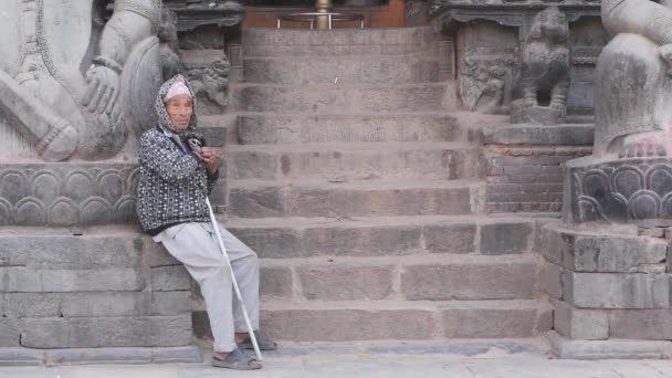 БХАКТАПУР, КАТХМАНДУ, НЕПАЛ - 18 октября 2018 года Бедняга на каменных ступеньках. Пожилой бедный человек сидит на пустых каменных ступенях индуистского храма. повседневная жизнь, восточный древний город после землетрясения — стоковое видео