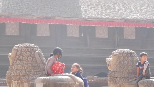 BHAKTAPUR, KATHMANDU, NEPAL - 18 Outubro 2018 Pequenas crianças alegres meninas sentadas em escultura de pedra, brincando no quintal do templo envelhecido à luz do sol. Vida diária, cidade antiga oriental após terremoto — Vídeo de Stock